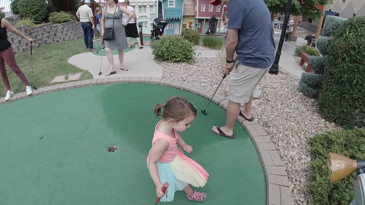 A young girl looks down toward the putting surface while adults with putters stand around the green and a blue ball sits near the hole area.