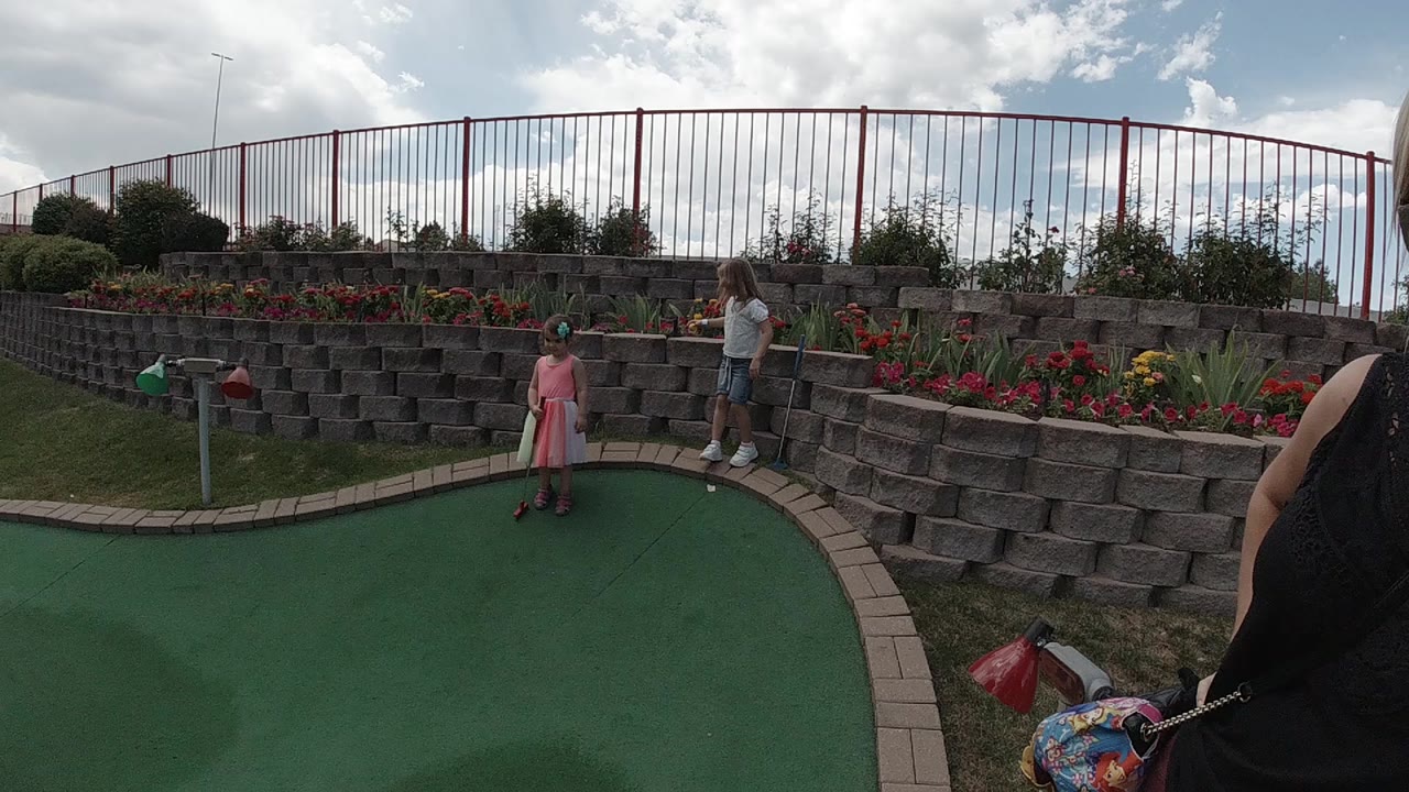 At 0:00, a girl in a pink dress stands on a Colorado mini golf green while an adult waits near a stone retaining wall and flower bed.