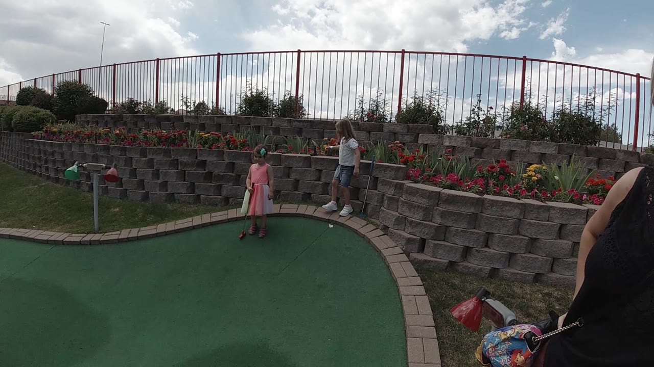 At 0:02, the girl stands slightly left of center on the green while the adult remains by the stone blocks under broken cloud cover.