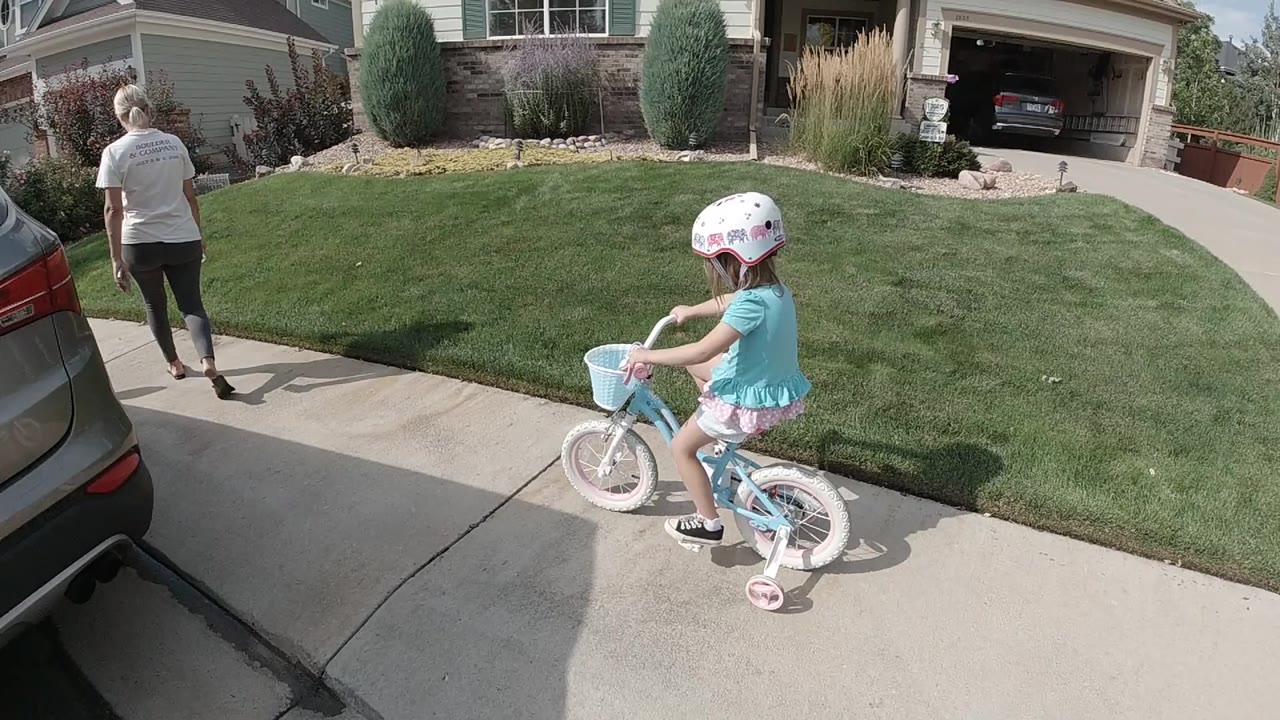 High-angle Colorado frame of a helmeted child starting along a concrete driveway on a small blue bicycle, with an adult walking ahead near a green lawn and parked cars.