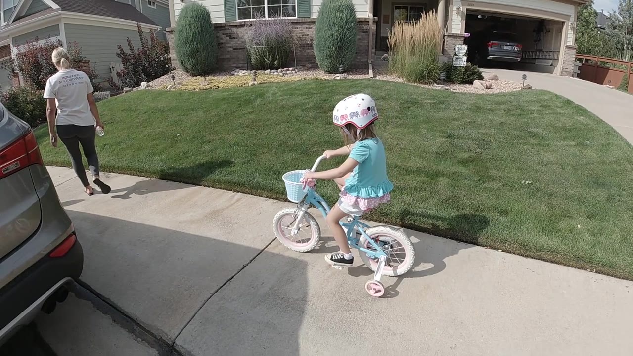 A high-angle view shows the child on the right side of the driveway, the adult ahead on the sidewalk, and the suburban Colorado lawn and house filling the upper frame.