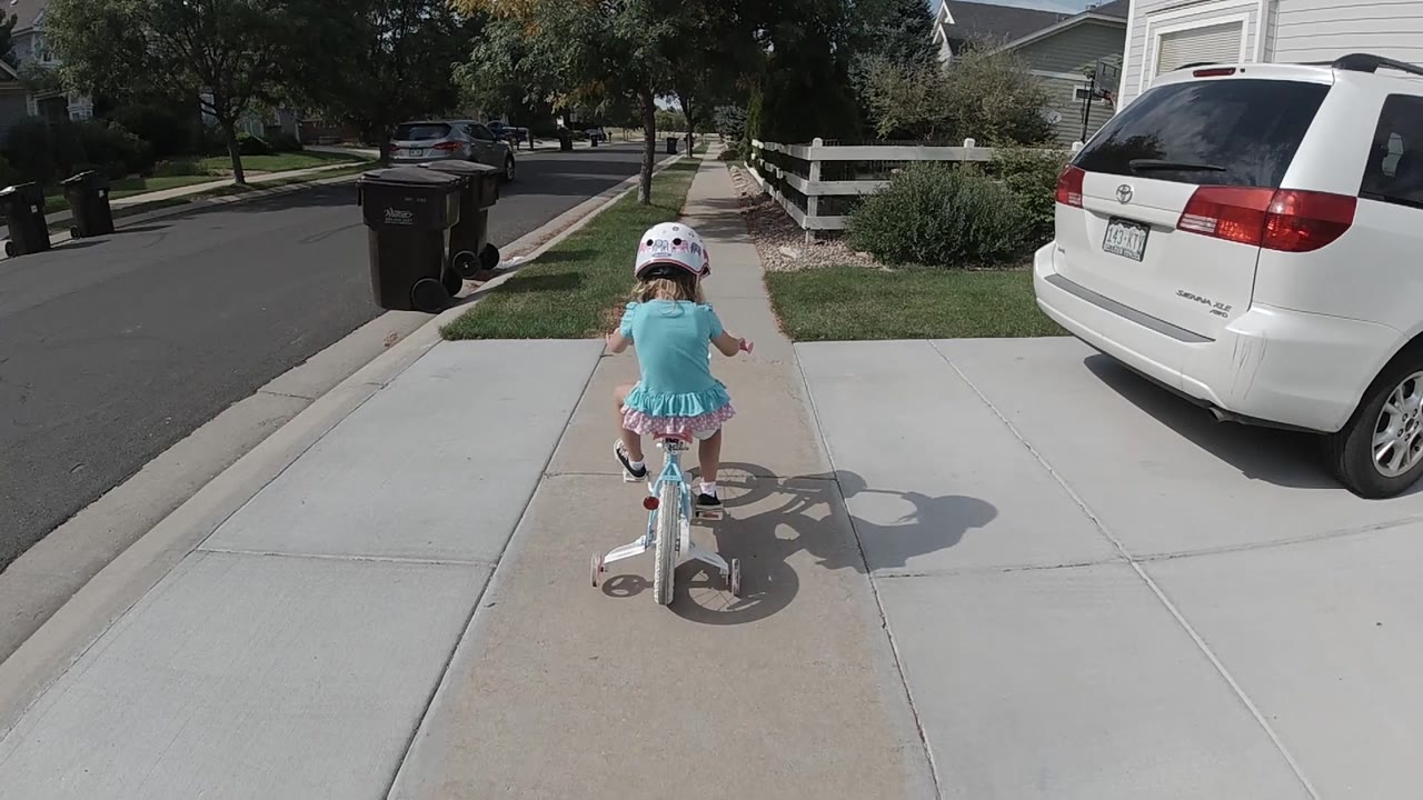 The toddler rides farther along the sidewalk, with the curbside trash bin ahead left, mowed grass on both sides, and the parked minivan along the driveway edge.
