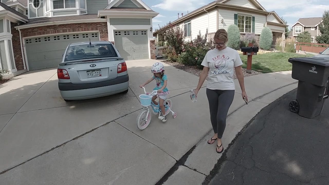 In Colorado, a helmeted child sits on a small blue bicycle with training wheels while an adult woman walks beside the driveway near a parked sedan and garage.