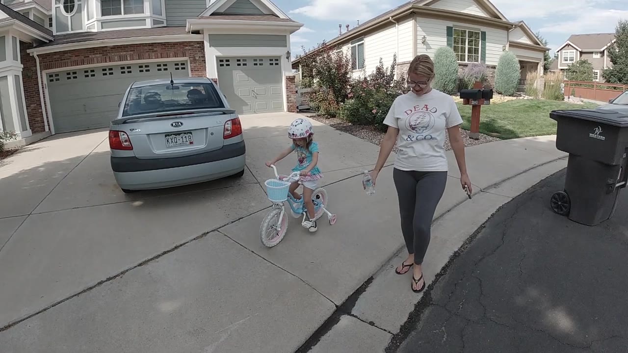 The child begins moving along the driveway on the training-wheel bicycle as the adult walks a few steps to the right of the bike near the street edge.