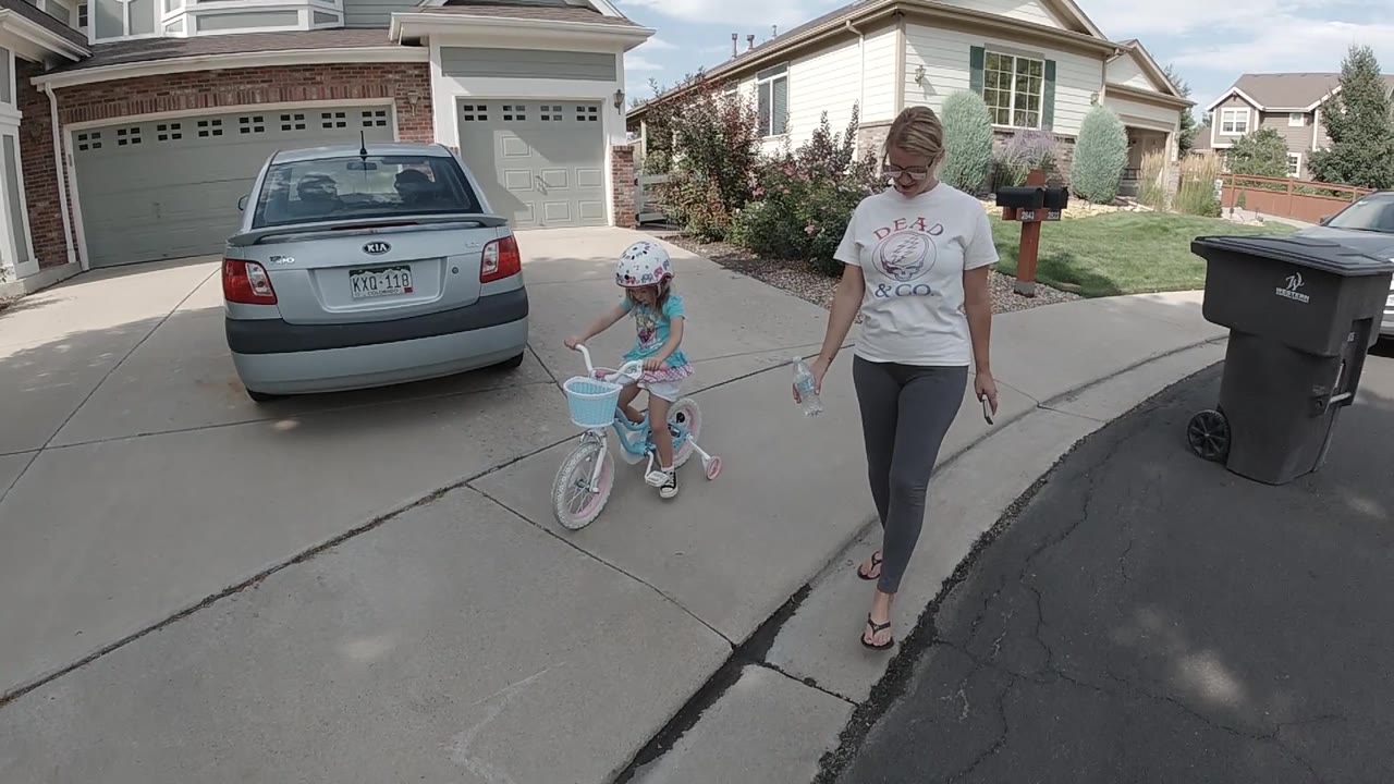 The child rides forward on the concrete driveway while the adult keeps pace beside the bike, with the parked sedan and double garage filling the background.