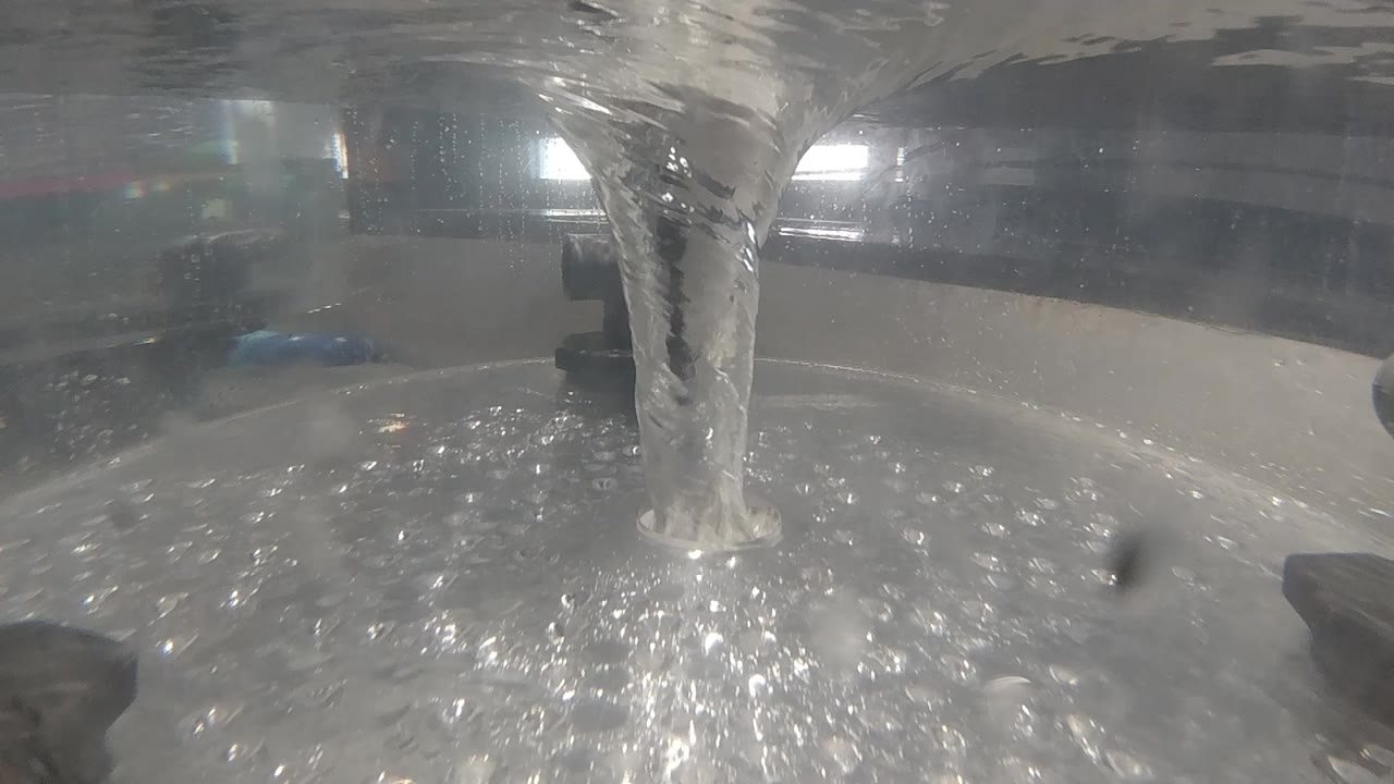 A continuous vertical water stream pours into a thermal pool in Colorado; rippling silver water surface fills the frame with two diffused light orbs visible through mist above; no visible people.