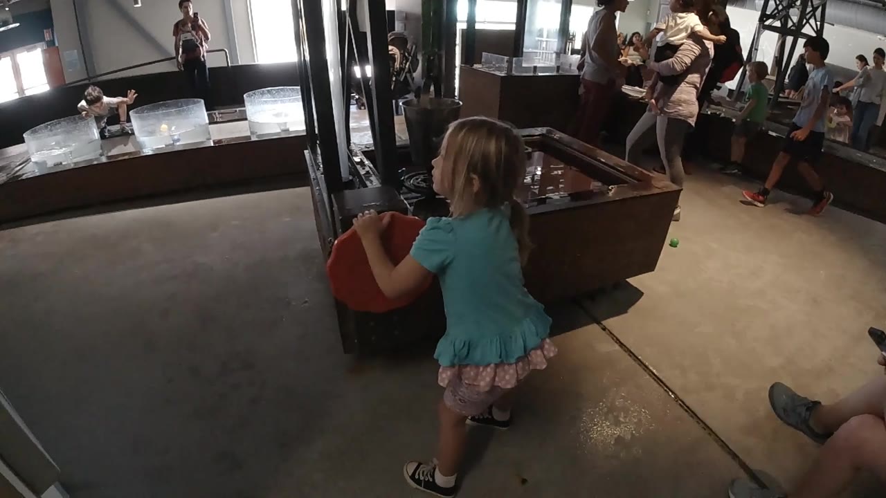 At 0:02, the toddler faces the dark structure while holding the red object, with seated and standing visitors visible across the room.