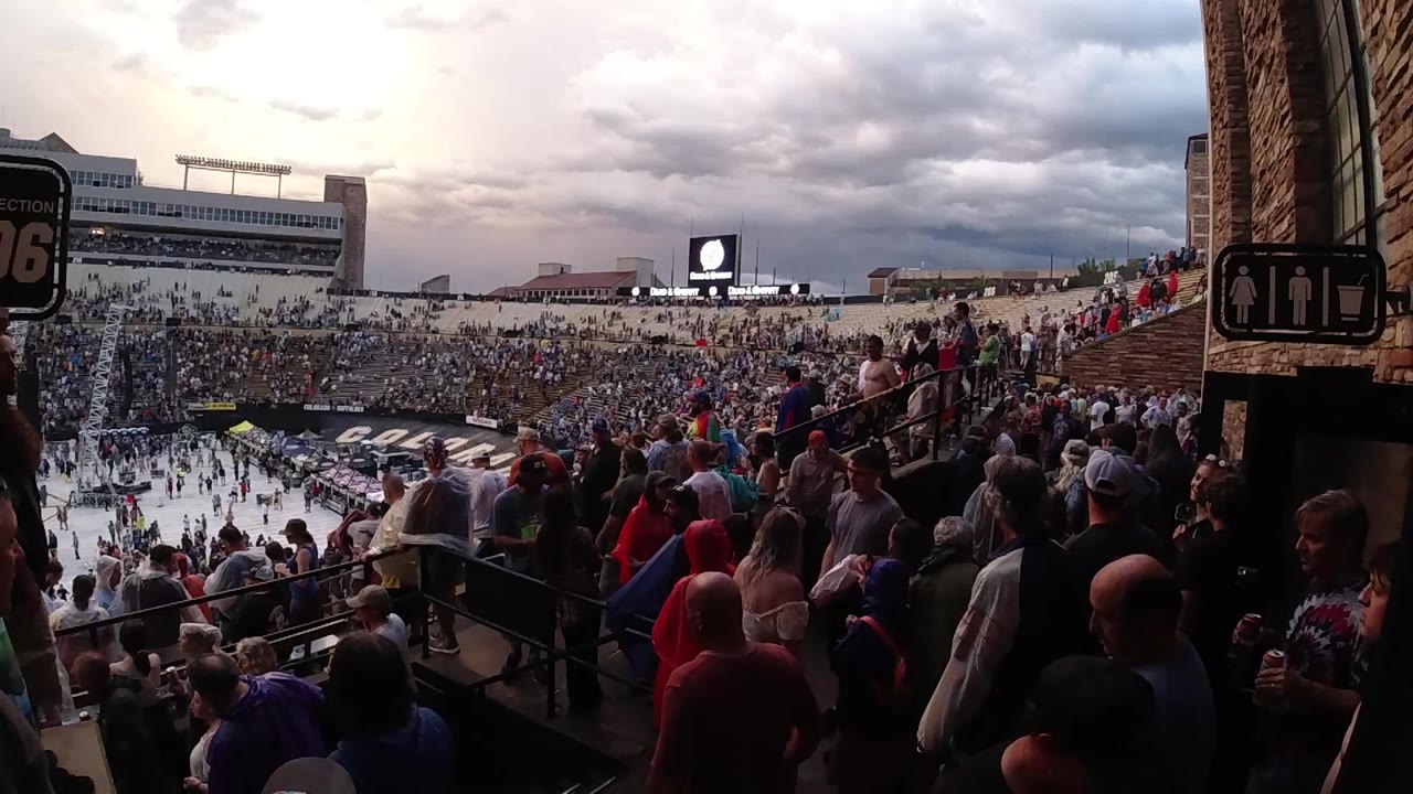 Wide Colorado stadium view from the bleachers, with a dense crowd in the foreground and a distant video screen under heavy clouds.