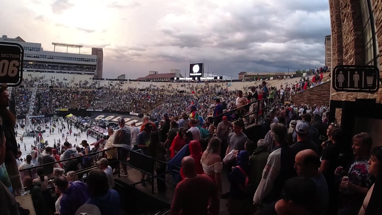 Wide bleacher view in a Colorado stadium, with spectators clustered along the aisle and a bright video screen beyond the field.