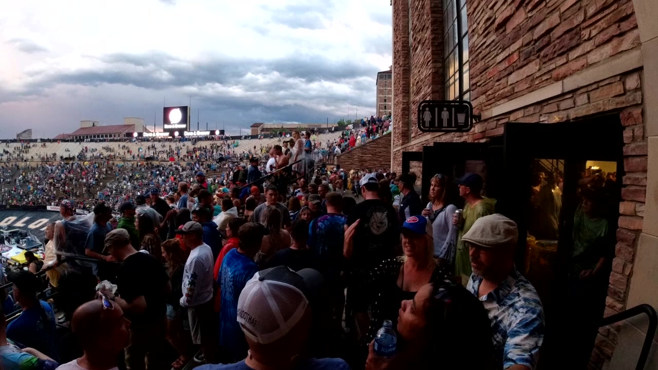 A Colorado crowd fills an outdoor concourse beside a brick building entrance, with a bright distant screen and overcast sky in the background.