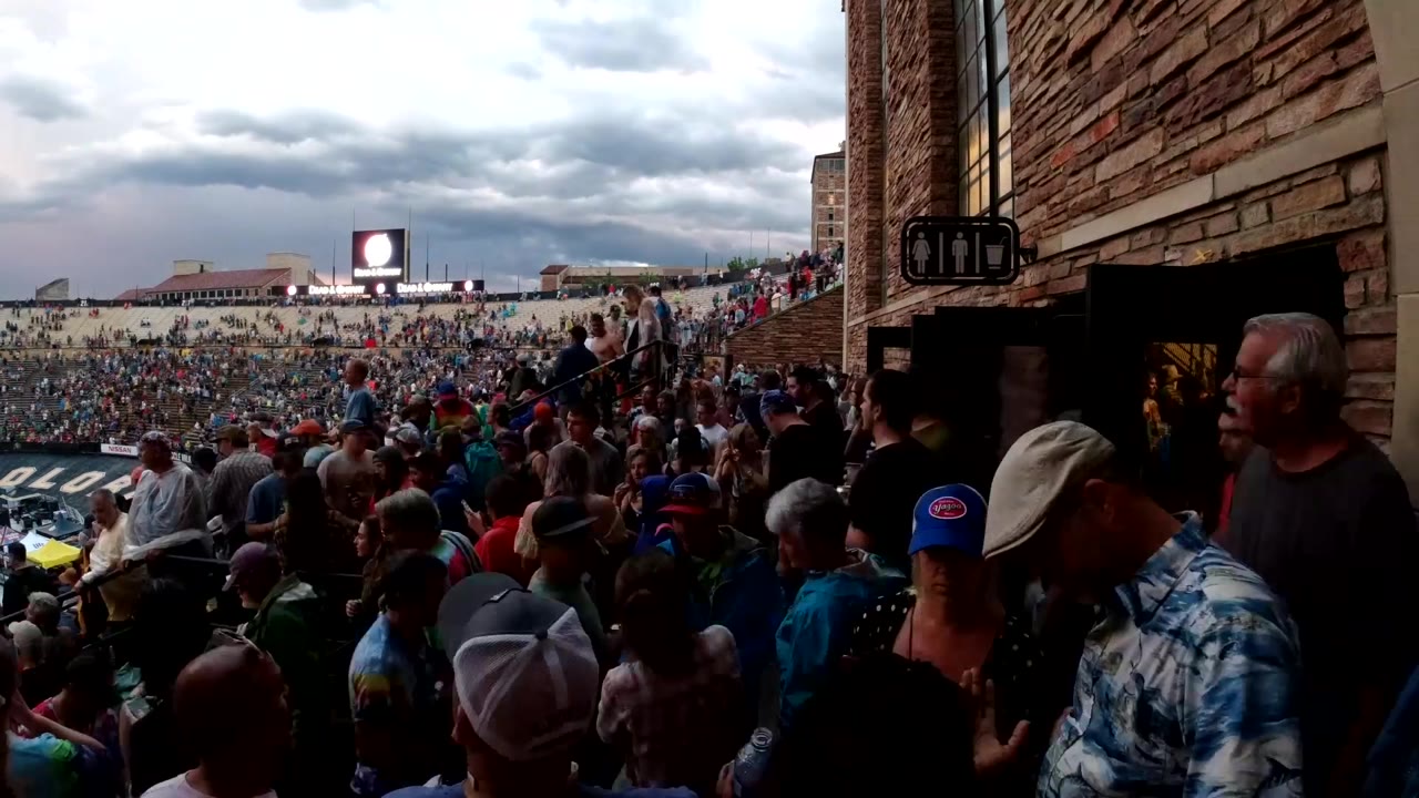 A dense Colorado event crowd stretches across the background while foreground attendees stand beside the brick venue wall and entrance.