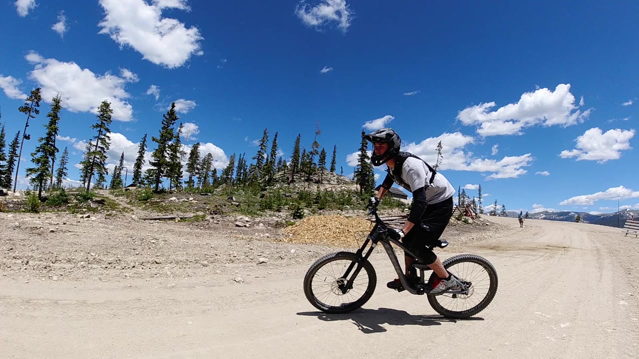 A mountain biker in helmet and backpack rides across a dusty Colorado forest road, with pine trees and bright clouds behind them.