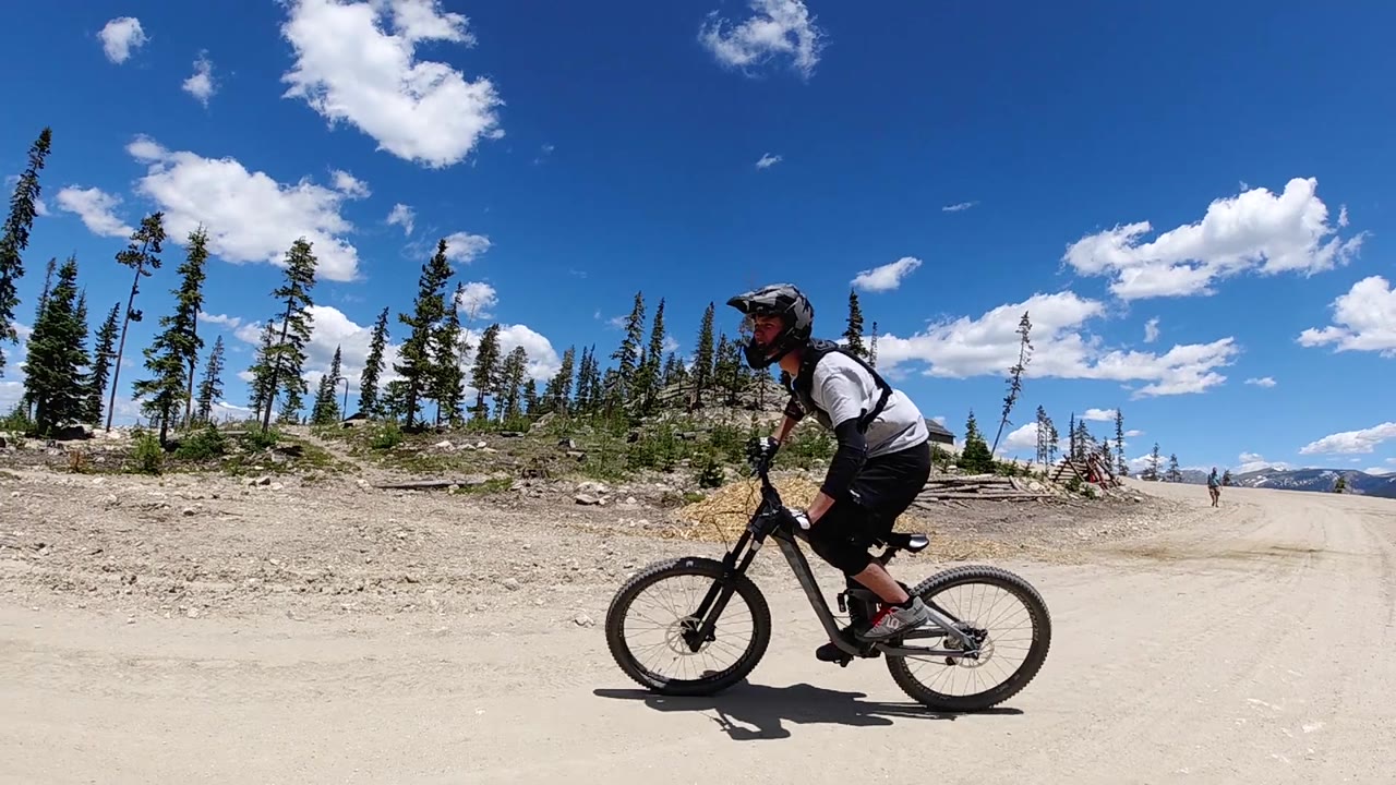 A mountain biker pedals through the right side of a broad dirt road, with thin pines and distant mountain shapes under a clear Colorado sky.