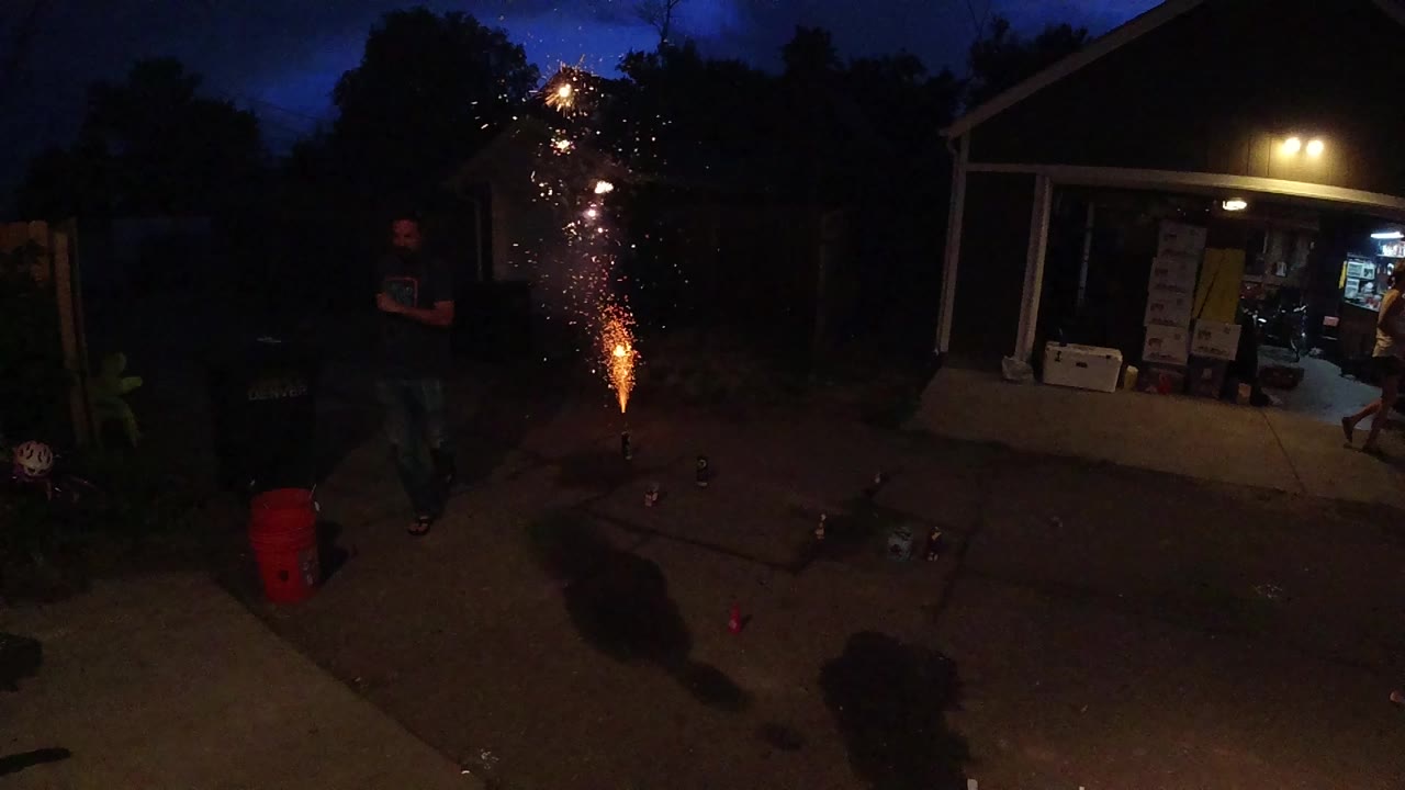 A Colorado backyard at night shows a ground firework throwing orange sparks near a lit porch, with an adult man standing in the darker foreground.