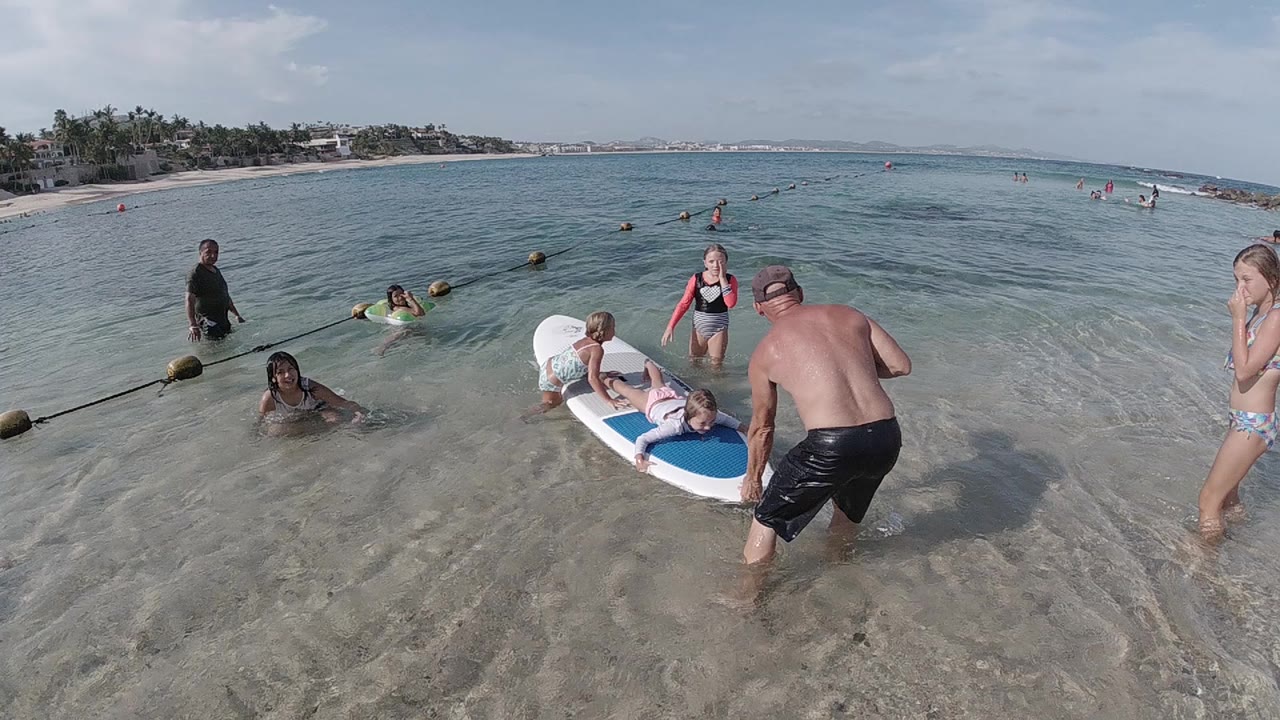 In Cabo, Baja California Sur, beachgoers stand and swim around a white and blue surfboard in clear shallow water as a shirtless man steps toward it.