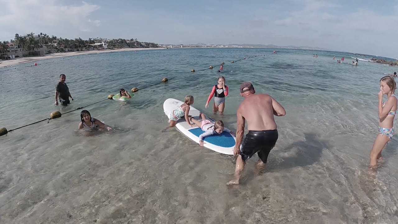 The surfboard floats in shallow Cabo water with children on top, a man facing it, a woman standing at frame right, and swimmers spread across the background.