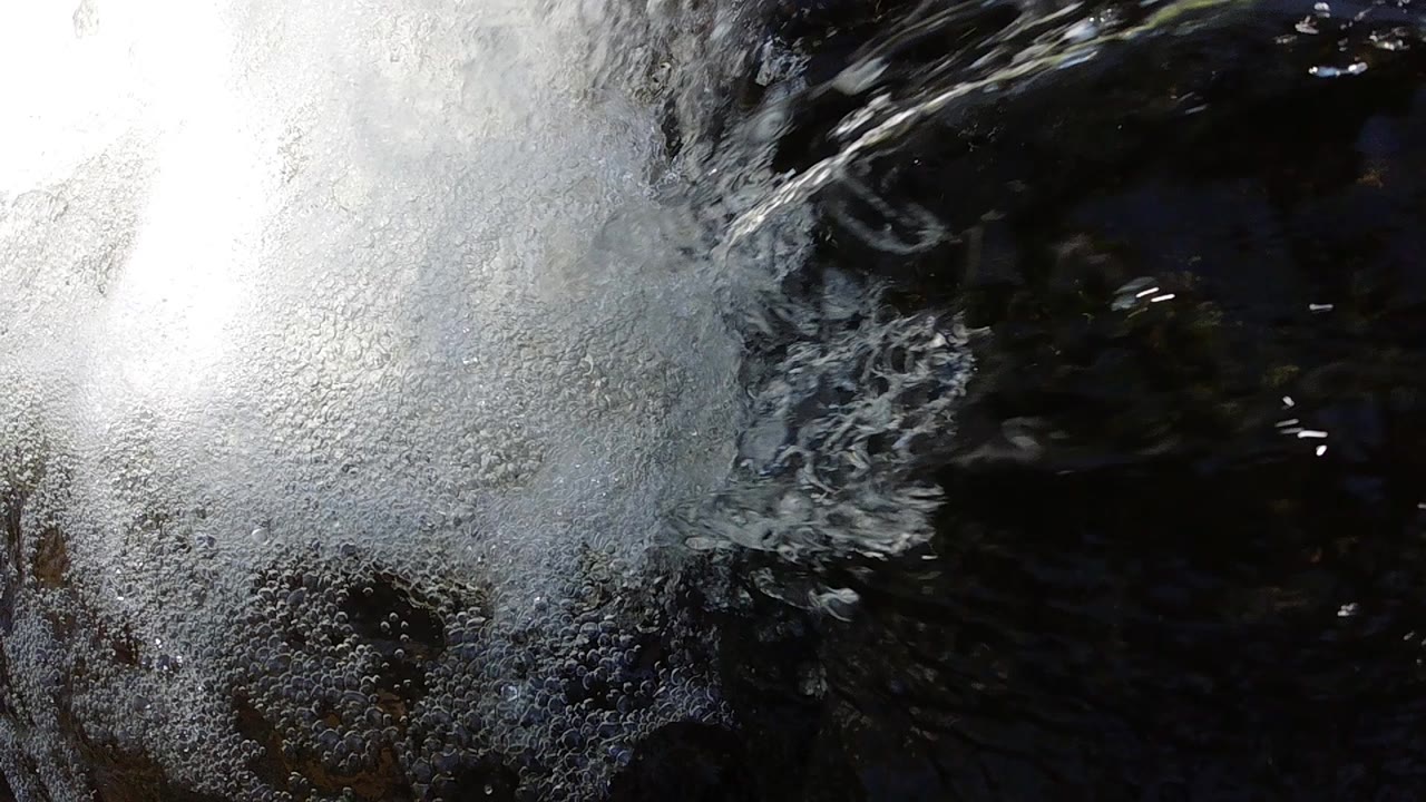 High-angle close-up aerial shot of bright white churning foam where a waterfall impacts a dark shadowed pool in Colorado; no people visible