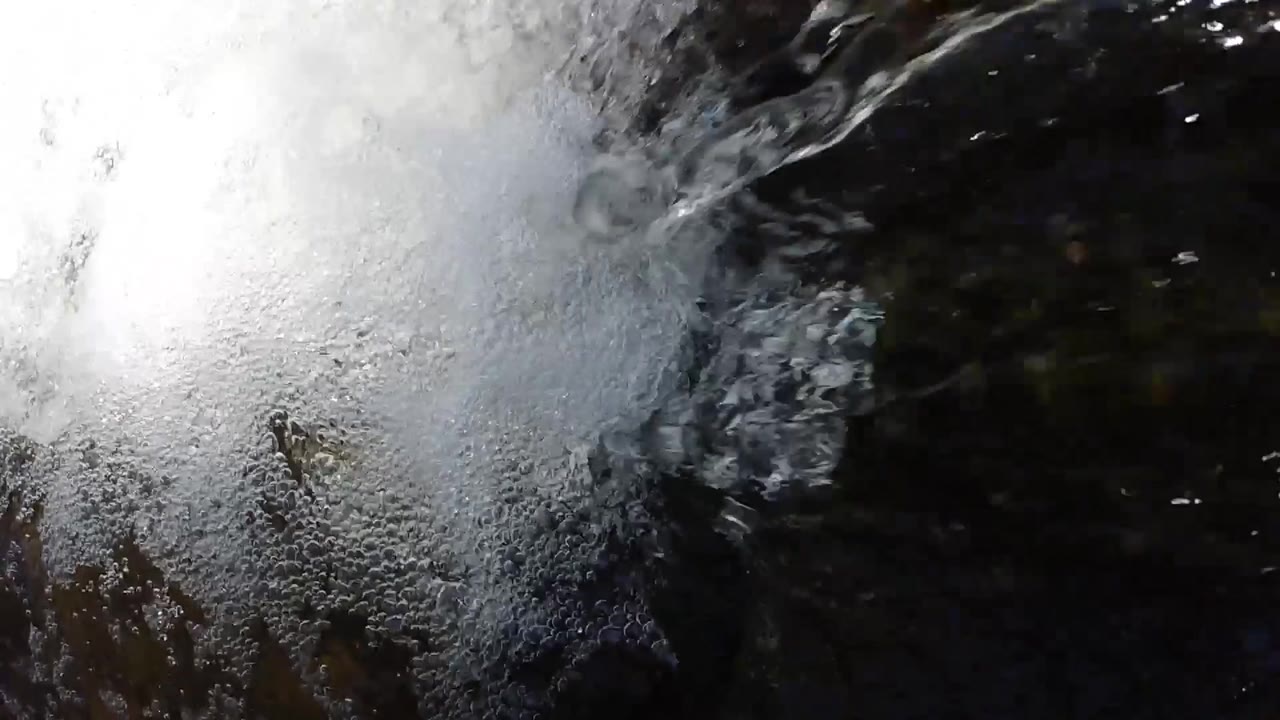 Close-up aerial shot of white churning foam from a waterfall impact zone with dark shadowed water in Colorado; no people visible
