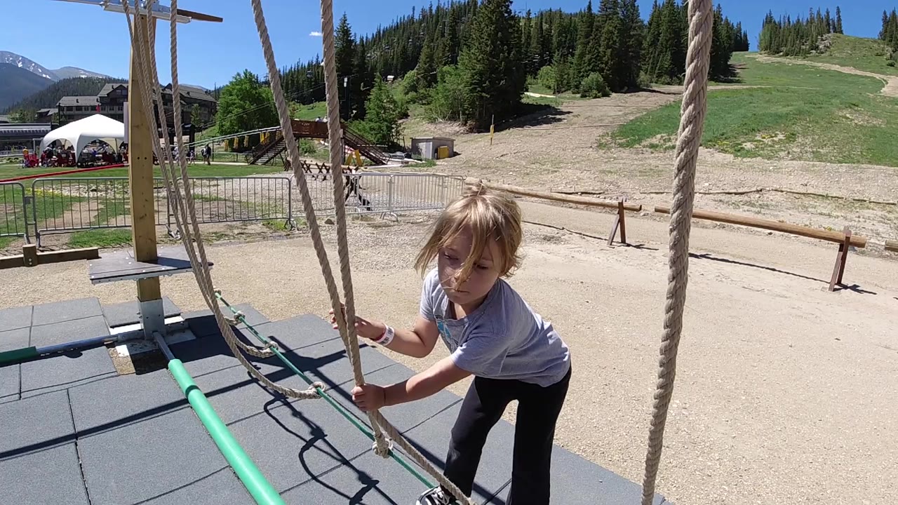 A young blonde girl in Colorado holds two rope lines near the start of an outdoor rope obstacle course, with rubber mats and a green pipe below her.