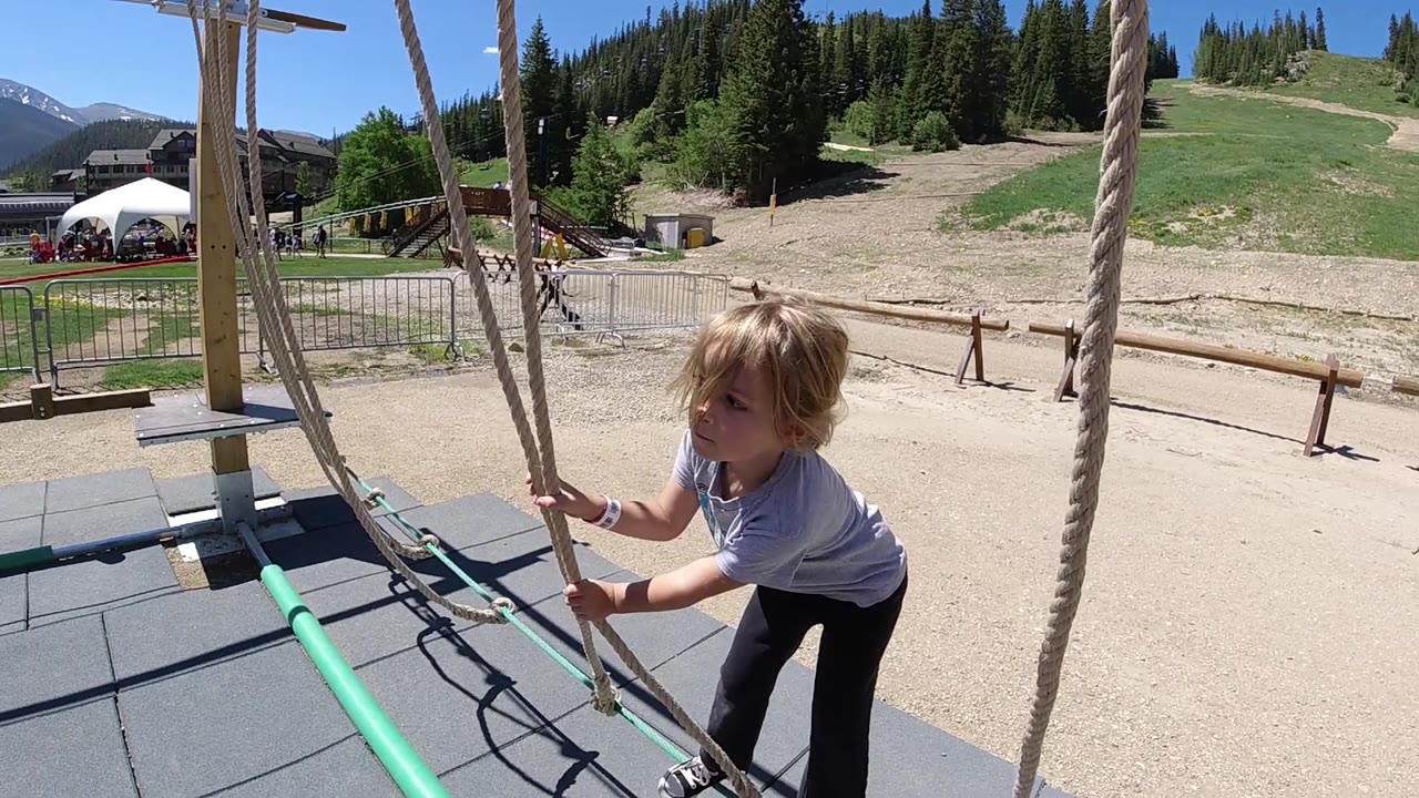 The girl looks to the side while keeping both hands on the rope lines, framed by vertical ropes and a sunny hillside beyond the obstacle course.