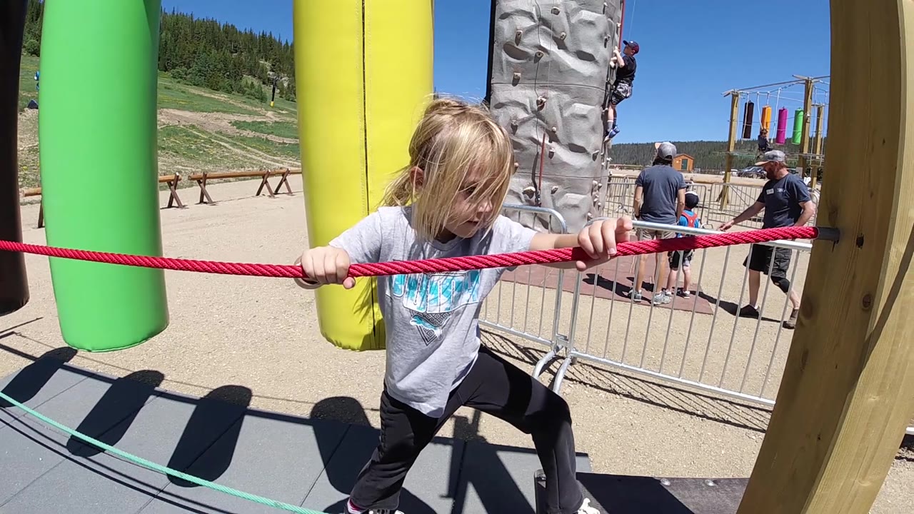 A young girl pulls the red rope while standing beside a yellow inflatable obstacle, with fencing and adults visible behind the Colorado playground course.