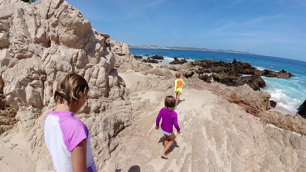 At 0:00, three children move away along a pale rocky coastal path in Cabo, Baja California Sur, with the nearest child in pink and white at left and surf breaking to the right.