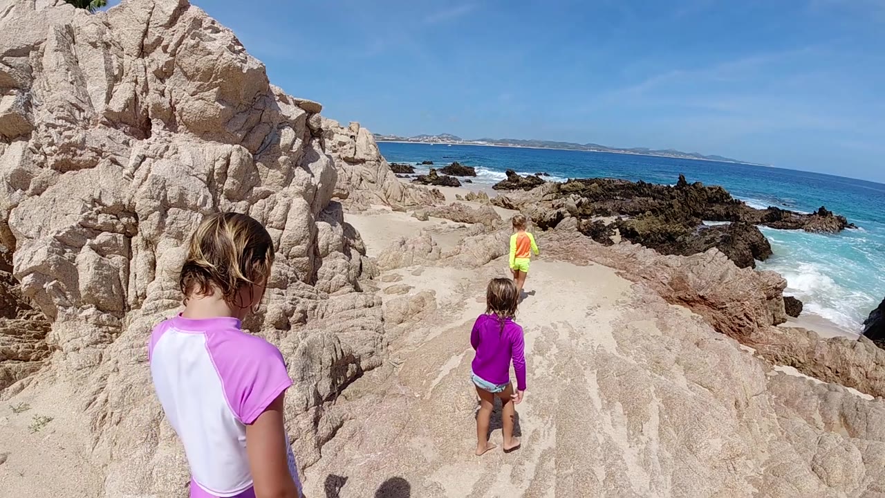 At 0:02, the nearest child in a pink and white suit stands near the left edge as two children ahead continue along the sunlit rock path toward the ocean horizon in Cabo.