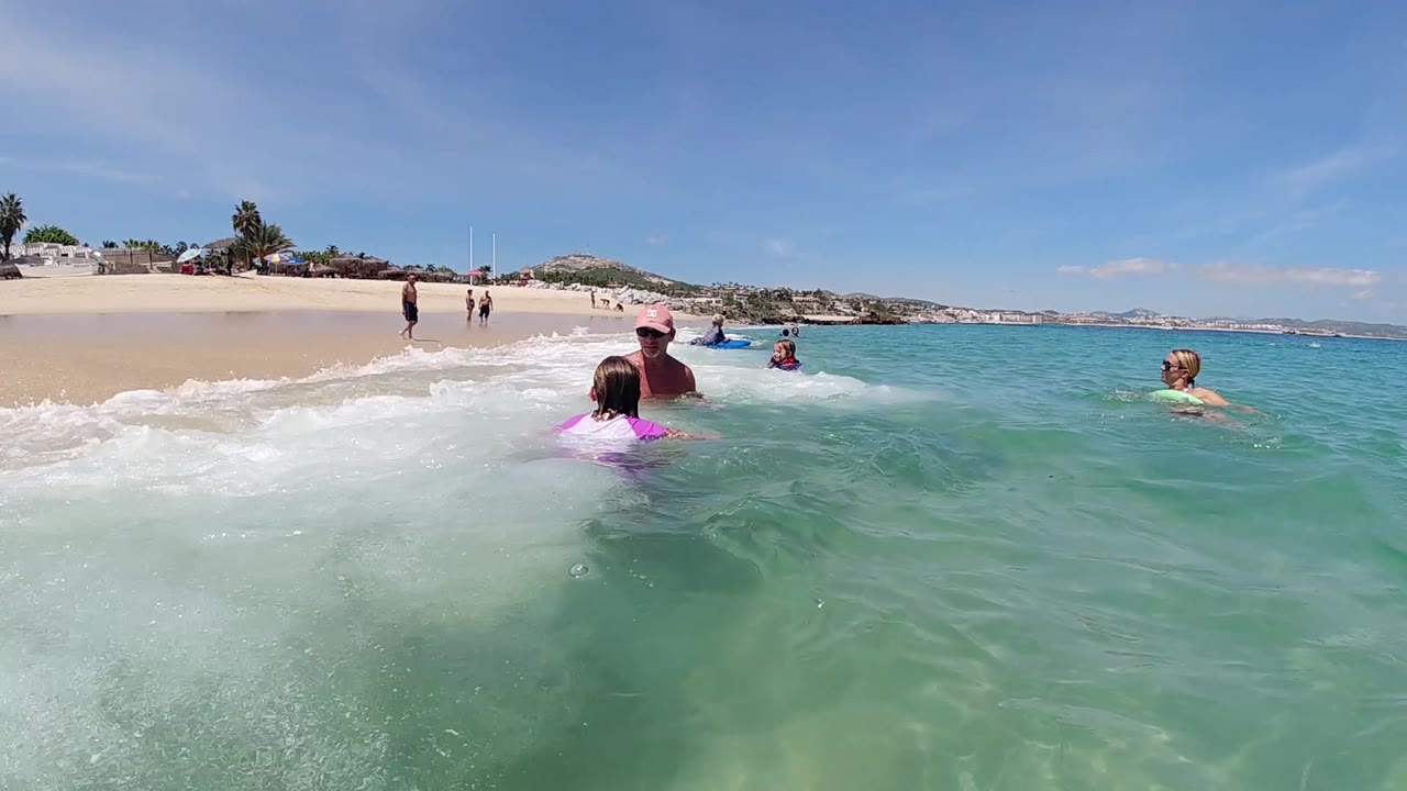 At 0:02, the child floats toward the adult in shallow water as a swimmer on the right and several beachgoers remain visible along the Cabo shoreline.