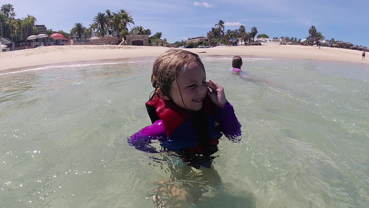 A toddler in a purple wetsuit and red-blue life vest faces the camera in clear shallow water at a Cabo beach, with a swimmer and sandy shore behind.