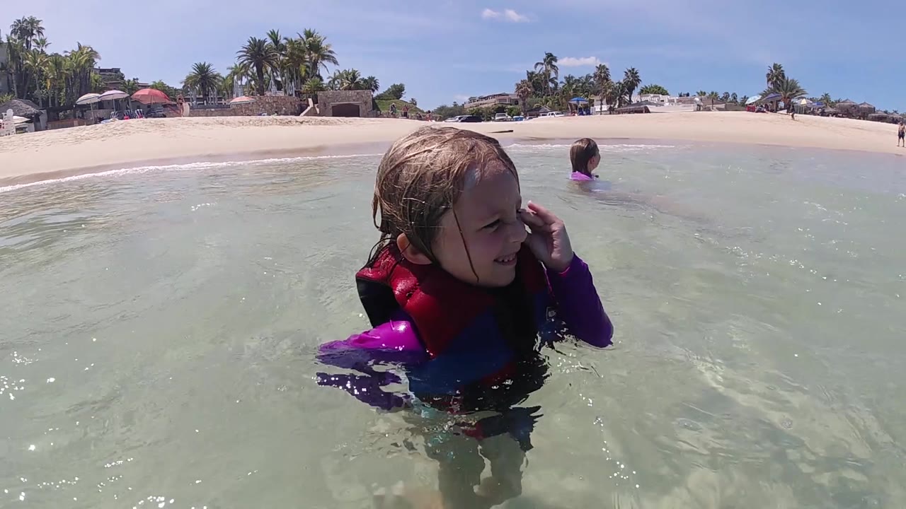 The child turns slightly to the right in waist-deep surf, with the sandy Cabo beach and another child farther back near the shoreline.