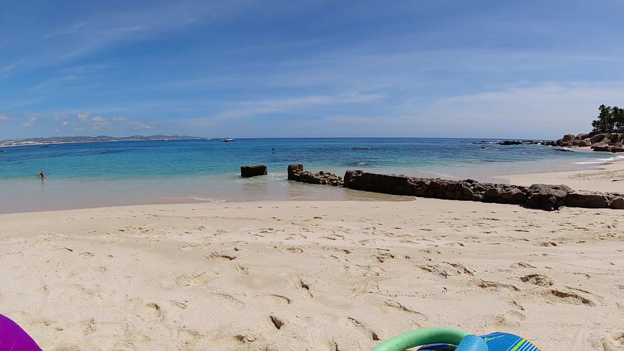 Ground-level frame from Cabo, Baja California Sur beach at one second, with a blue inflatable dominating the left foreground, a dark driftwood log crossing mid-frame, a magenta inflatable at the lower right, turquoise ocean on the horizon, and blue sky with wispy clouds; no visible people.