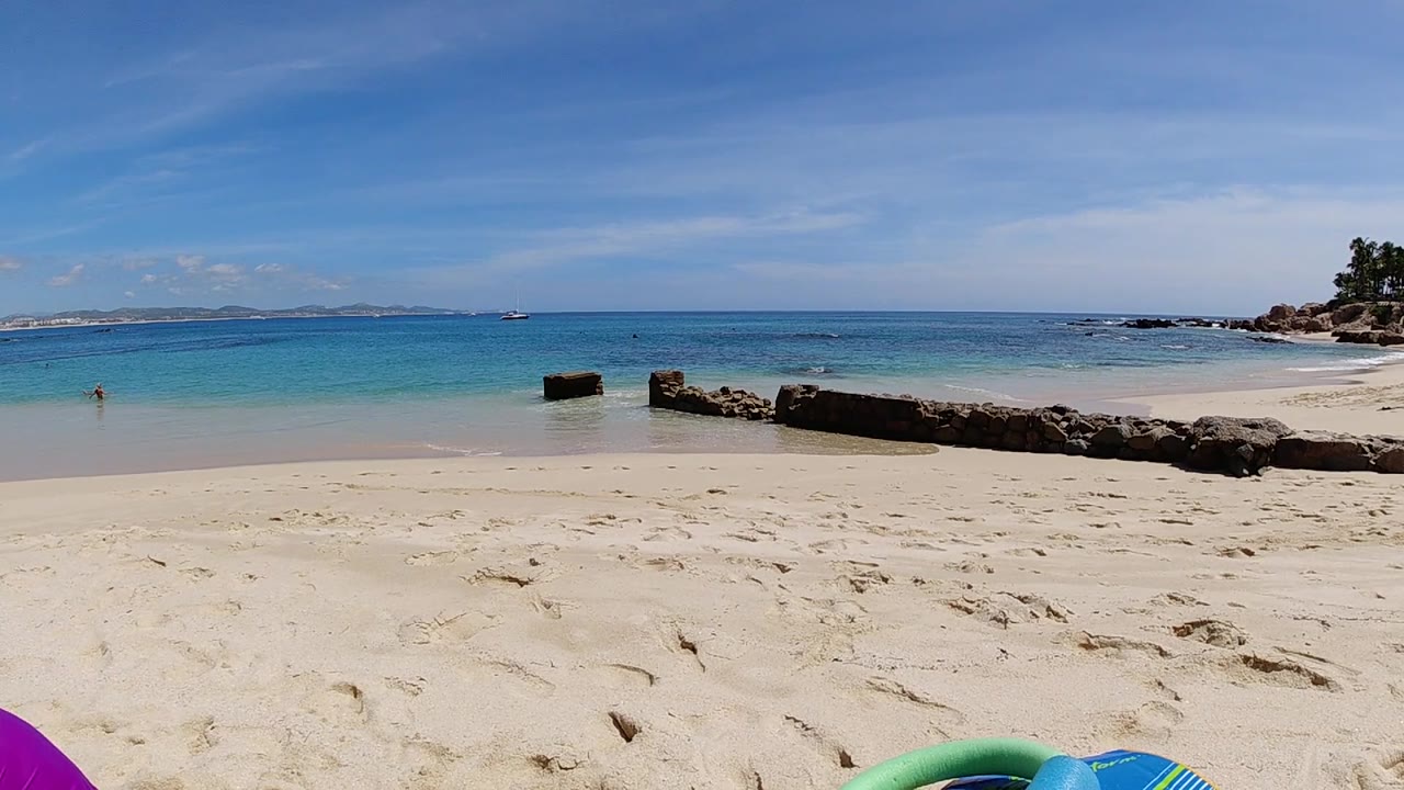 Ground-level beach frame from Cabo, Baja California Sur, Mexico at three seconds, with blue and green inflatables in the left foreground, a dark driftwood log mid-frame, magenta inflatable at the lower right, turquoise ocean, and blue sky; no visible people.