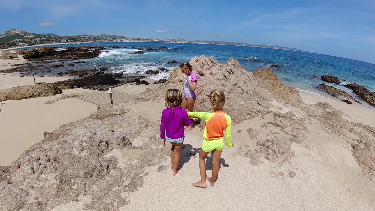 Children walk along pale sand ridge near rocky ocean shore.