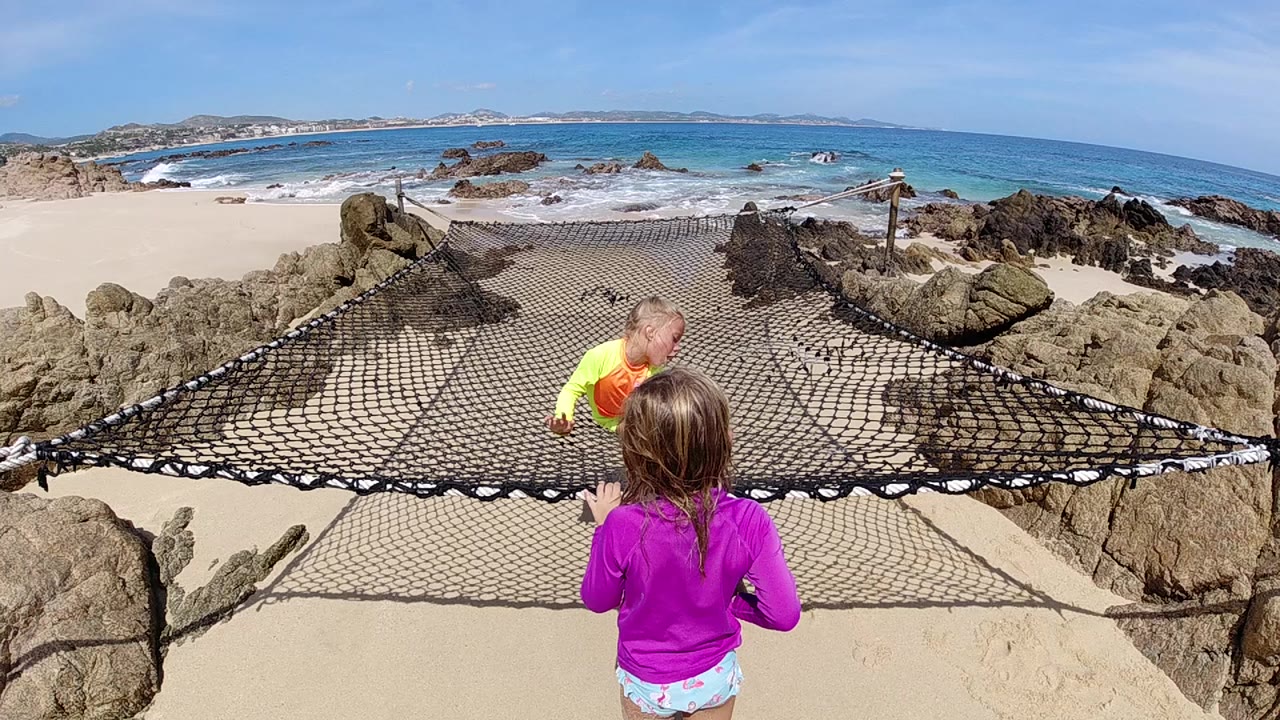 Two children play on a black rope safety net over sand in Cabo, Baja California Sur, with rocky edges, blue ocean, and a bright horizon behind them.