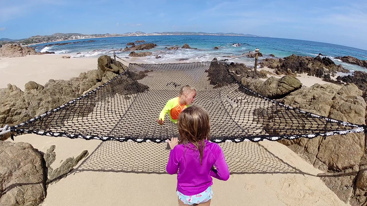 Girl in purple stands near a beach rope net while another child climbs.