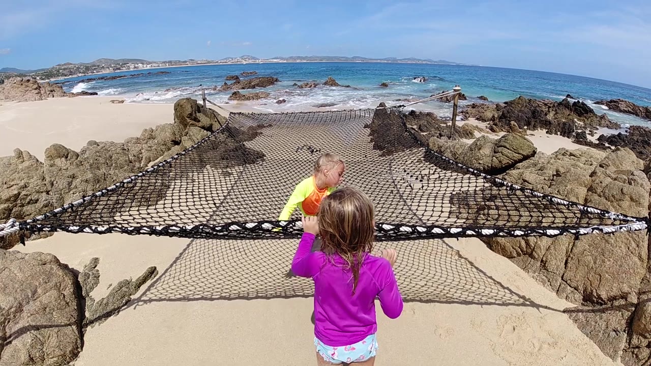 The climbing child reaches higher on the black rope net as the foreground girl raises a hand near the mesh, with pale sand and blue water beyond.