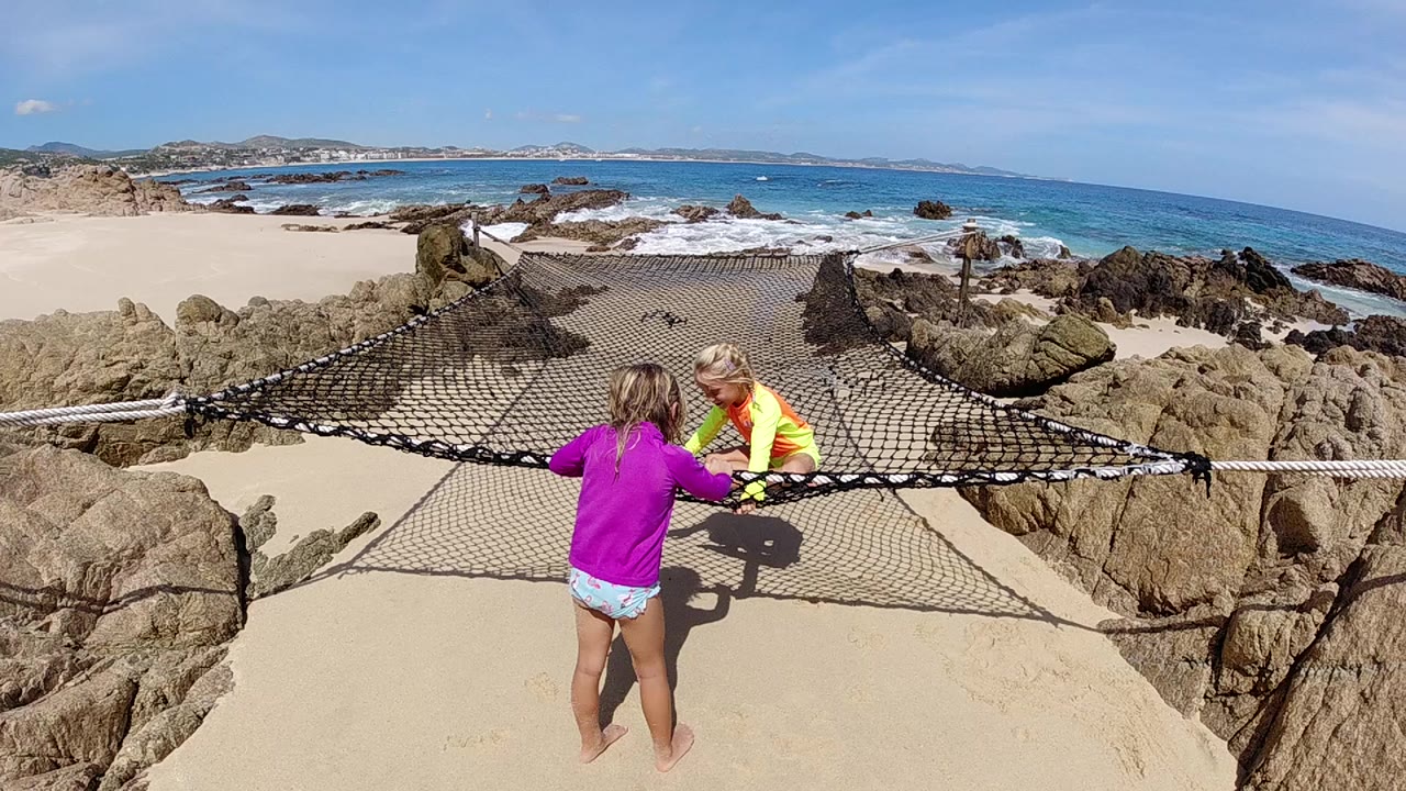 At the opening second in Cabo, a girl in a purple top faces a seated child in yellow on a rope net, with rocks, sand, and blue surf behind them.