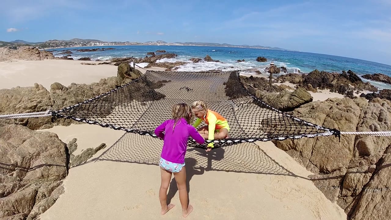 The girl remains at the lower center while the child in yellow sits on the net, framed by tan rocks and a bright blue Cabo shoreline.