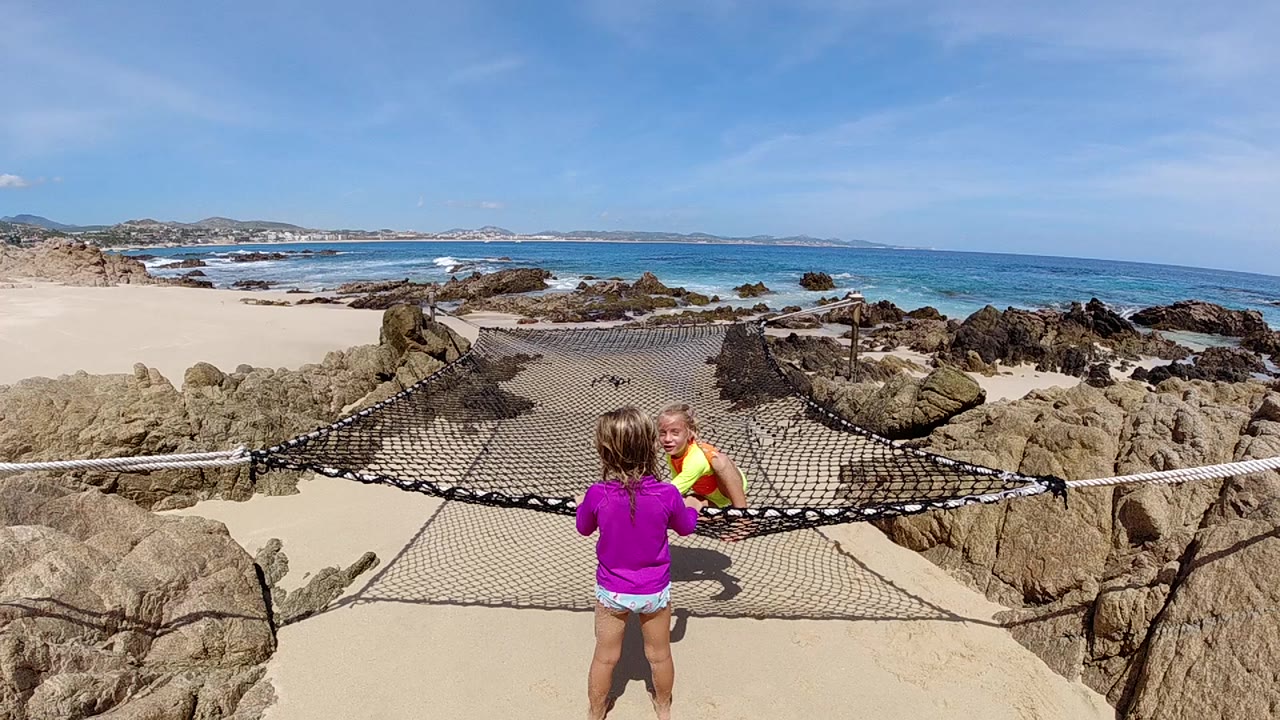 At the start, a girl in a purple shirt faces a boy in yellow beside a rope barrier on a rocky Cabo beach platform with blue ocean beyond.