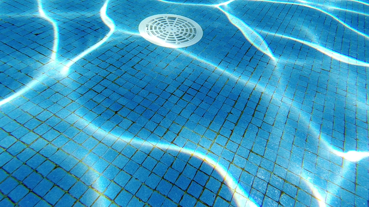 Blue tiled pool floor viewed from above through clear water, with a white circular drain cover and rippling caustic light patterns.