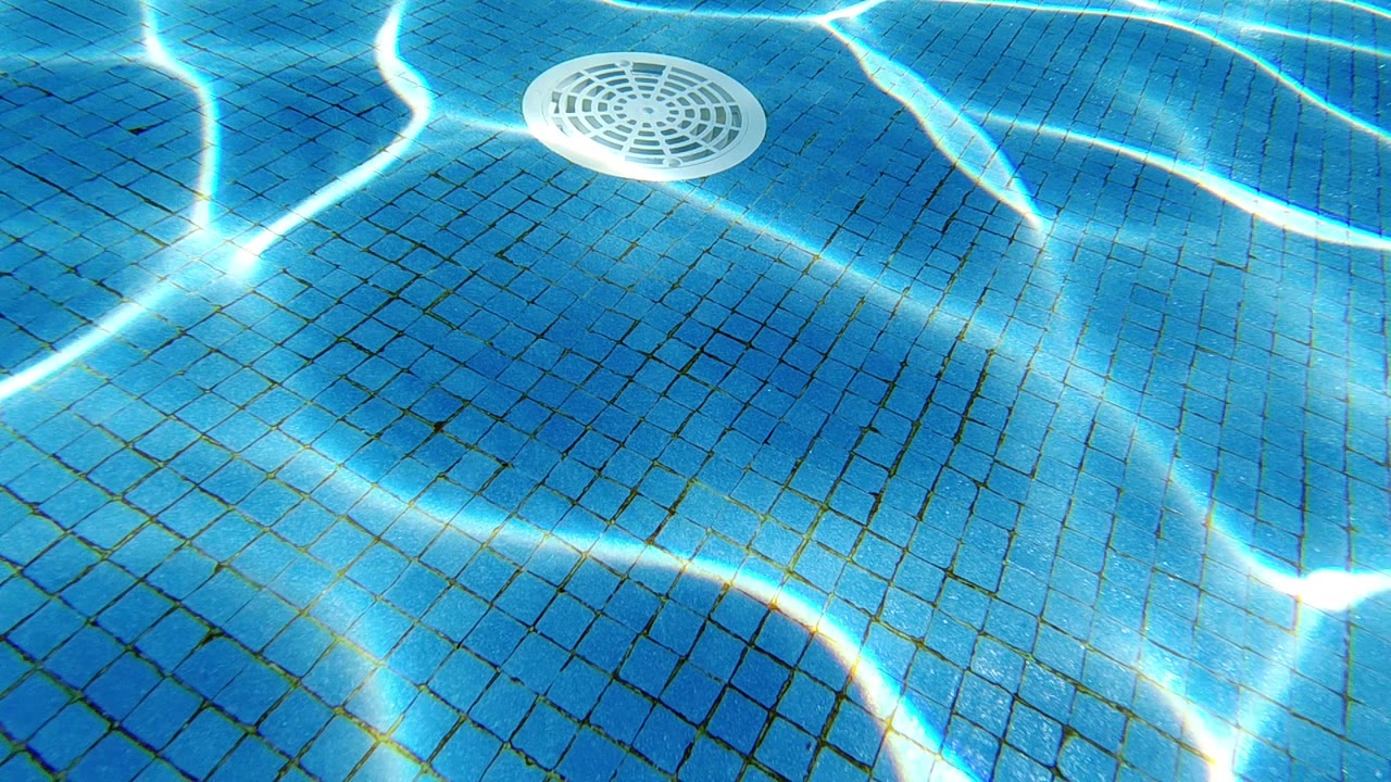 Overhead view of blue mosaic-tiled pool floor; white circular drain cover at center; bright caustic ripple patterns shifting across the tiles; no visible people.