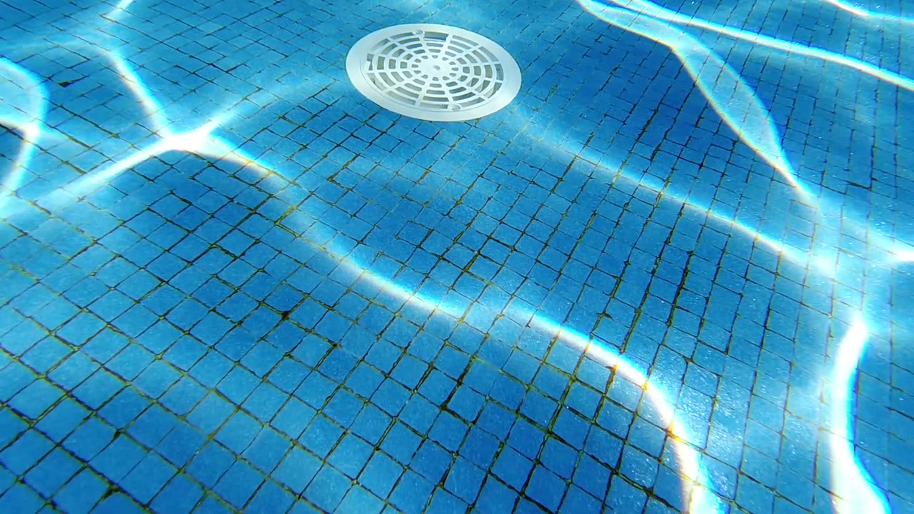 Overhead view of blue tiled pool floor; white circular drain cover shifted slightly right; bright caustic light patterns moving across the mosaic tiles; no visible people.