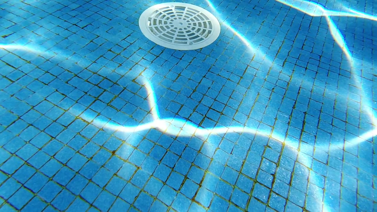 Overhead view of blue mosaic-tiled pool floor; white circular drain cover visible; bright caustic light patterns from sunlight refracted through the water surface; no visible people.