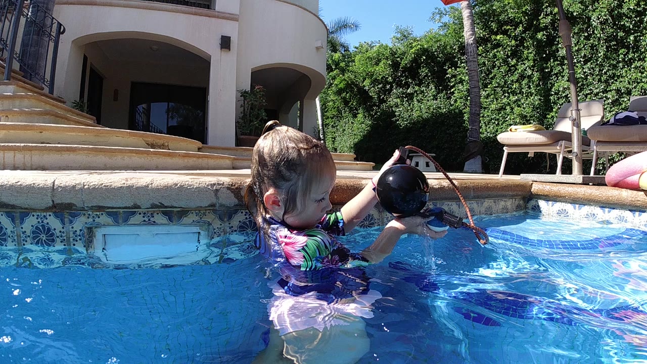 A toddler in a colorful swimsuit stands in a Cabo residential pool holding a dark toy above rippling blue water, with a white house and stairs behind.
