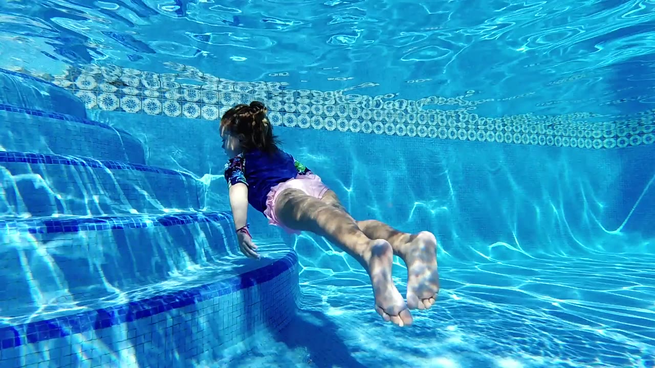 A girl glides underwater past the left-side steps, with her feet nearest the camera and the tiled pool wall behind her.