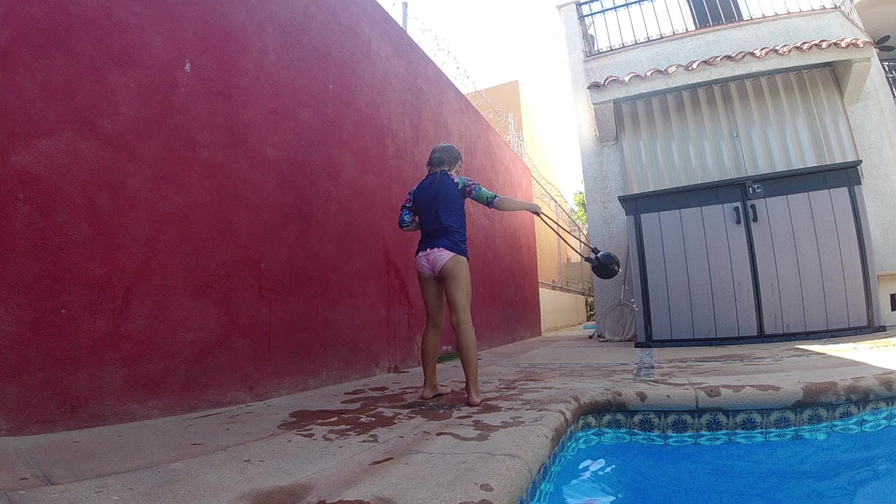 A woman in a blue top and pale shorts stands near the swimming pool in Cabo, holding a black skimmer toward the red courtyard wall.