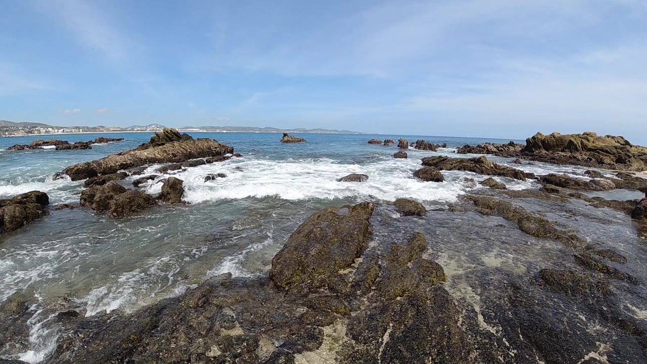 Low-altitude drone aerial over dark volcanic rocks along the Cabo Baja California Sur coastline, white sea foam churning through crevices, blue Pacific ocean extending to horizon under clear blue sky, no visible people