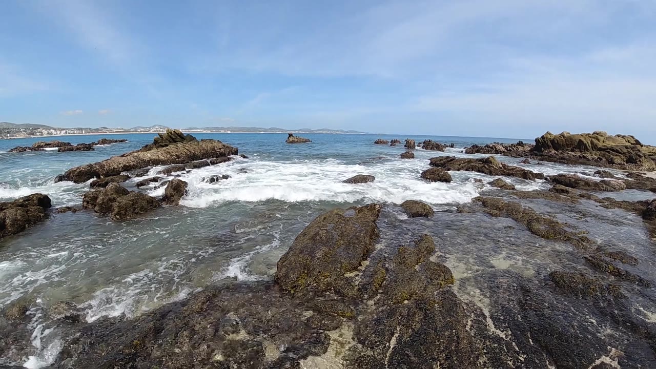 Low-altitude aerial over a dark volcanic rock platform on the Cabo Baja California Sur coastline, white foam active in the rock channels, blue Pacific ocean and clear blue sky in the background, no visible people
