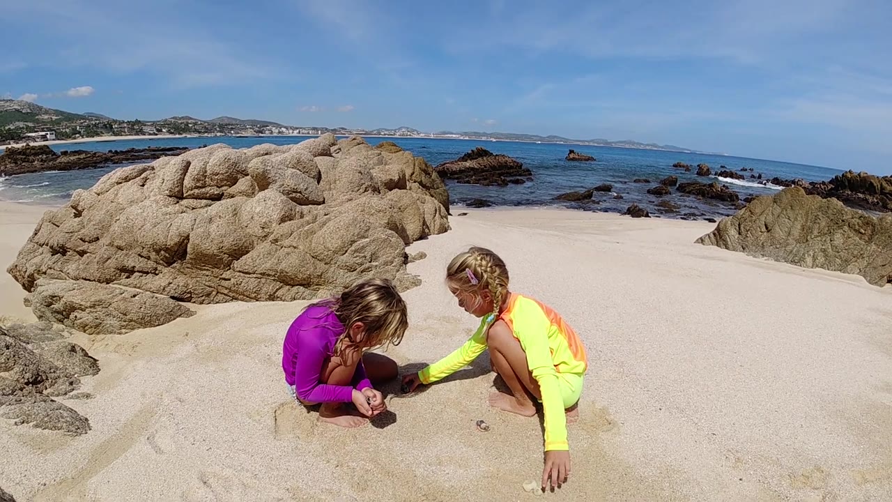 At 0:02, the children remain crouched on the sandy Cabo shore, framed below a large rounded rock with rippled ocean and dark rocks in the background.