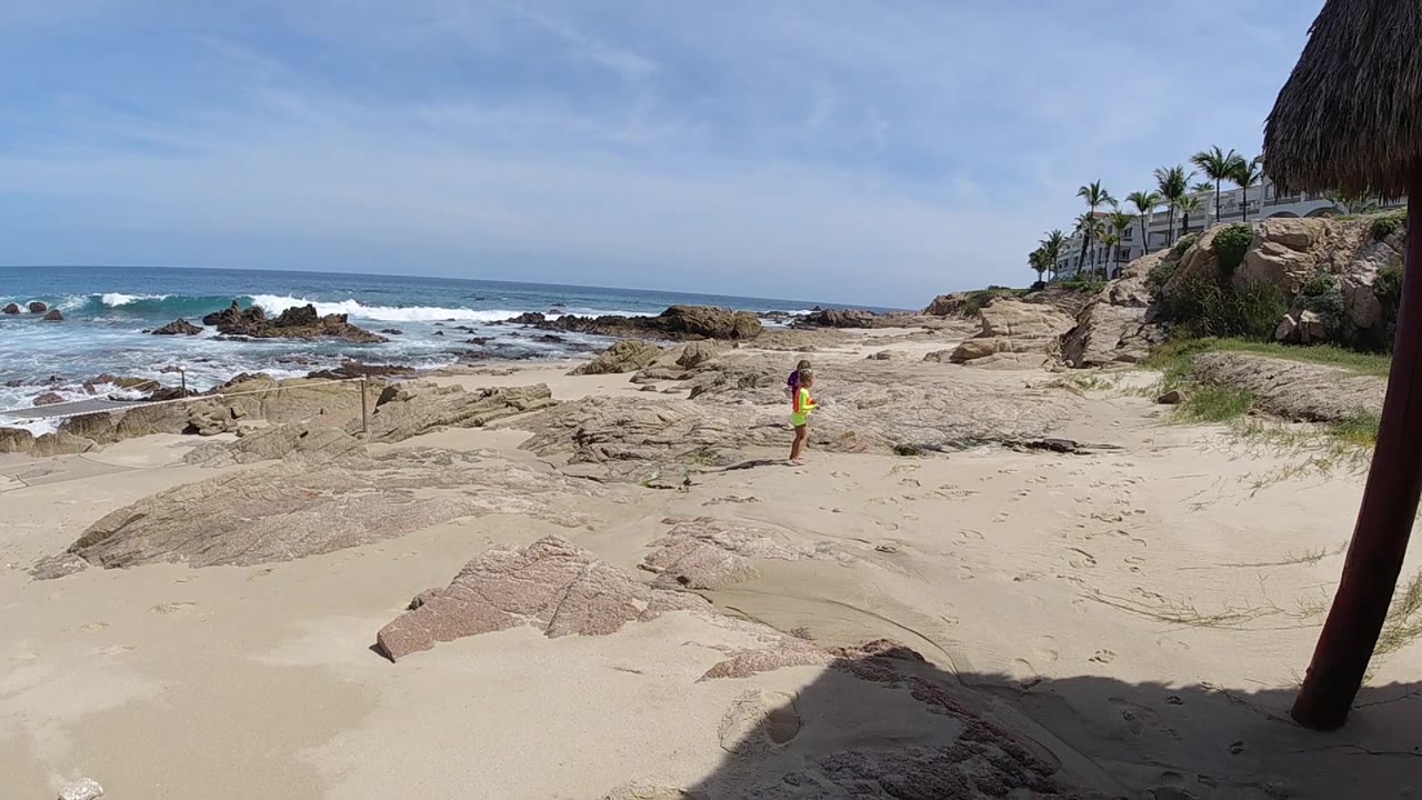 At 0:02 in Cabo, the lone person remains near center frame as the ocean breaks over dark rocks on the left and cliffside palms line the right background.